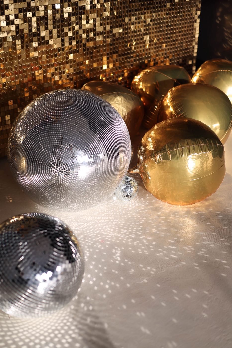 A gold sequin backdrop with gold balloons and mirror balls on the floor