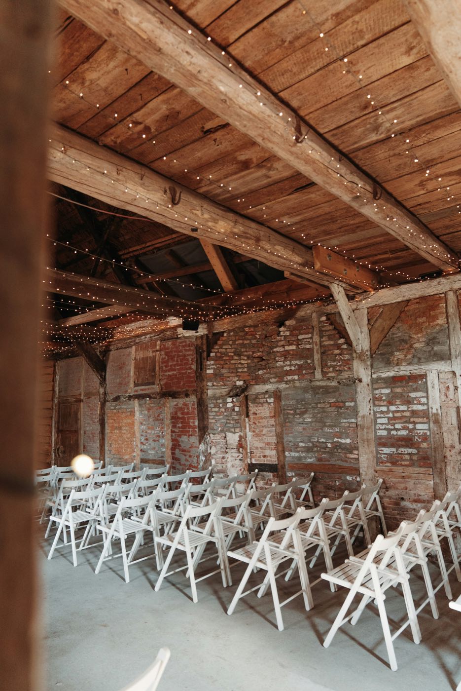 Rows of white folding chairs under string lights in a barn with brick walls and wooden beams