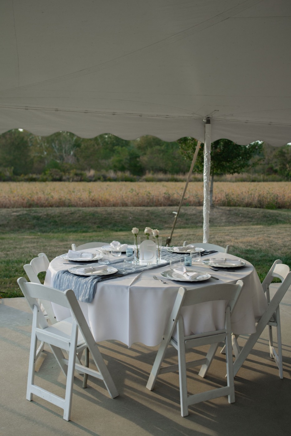 A round table in a white cloth with white folding chairs under the edge of a tent, with a field beyond