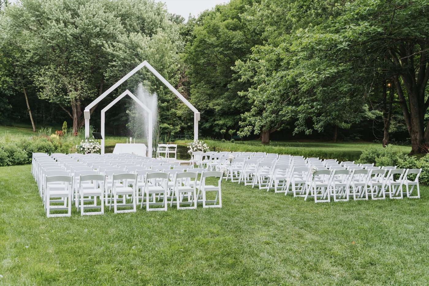 Rows of white folding chairs on a lawn facing a white arch, with tall trees behind