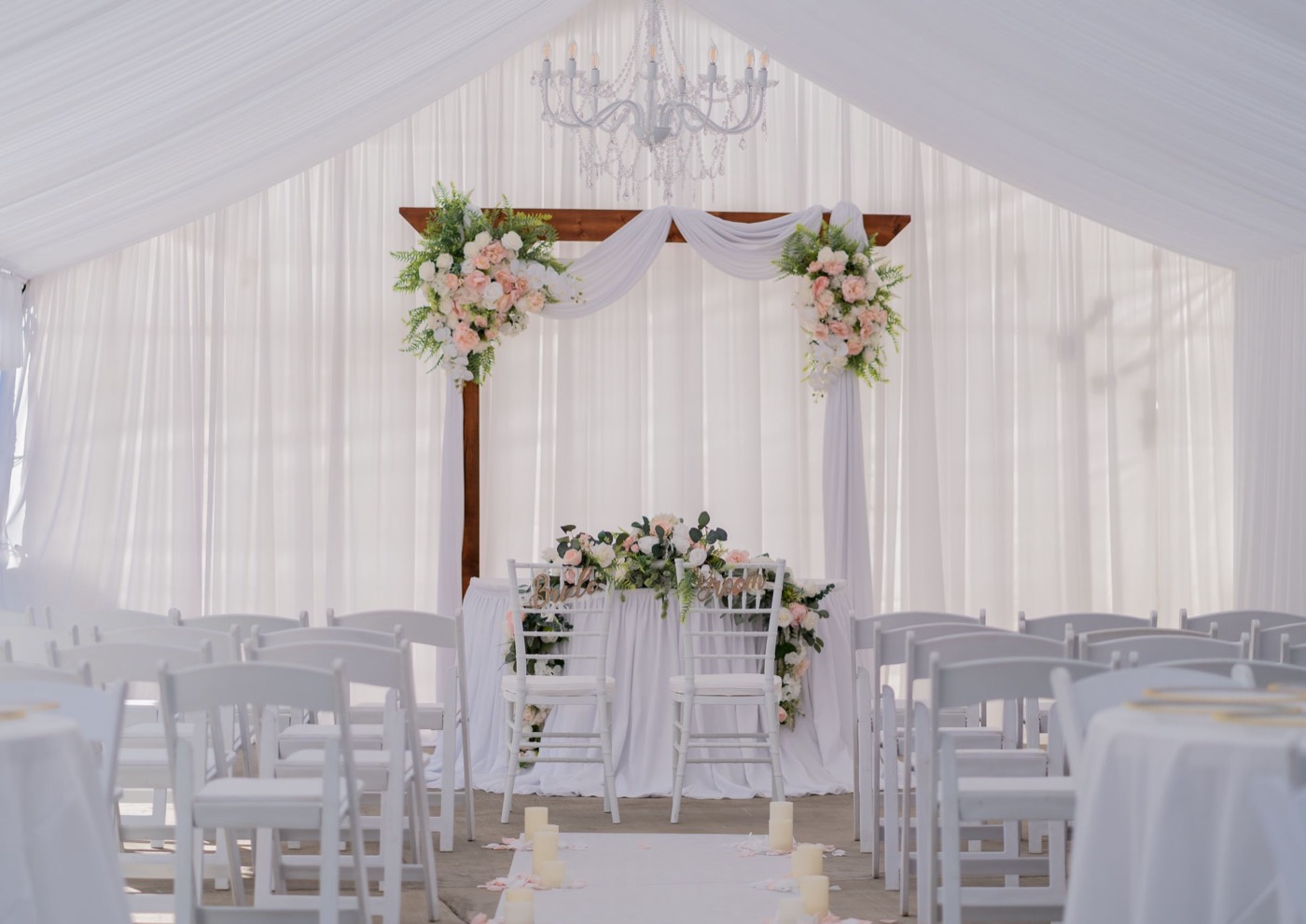 A draped white tent with a chandelier over a flowered arch and rows of white chairs