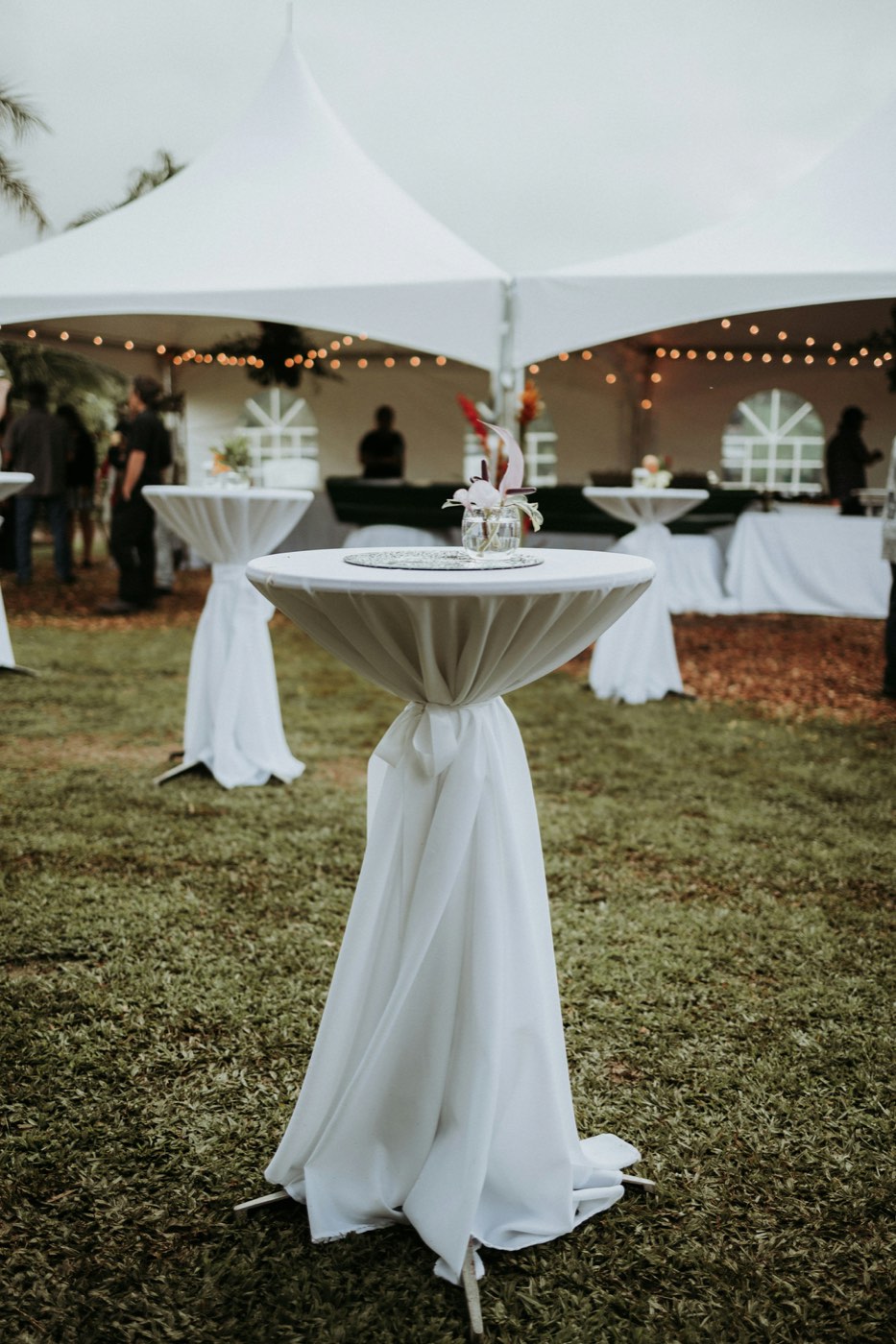 A white peaked frame tent with string lights on a lawn, with high-top tables in white linens in front