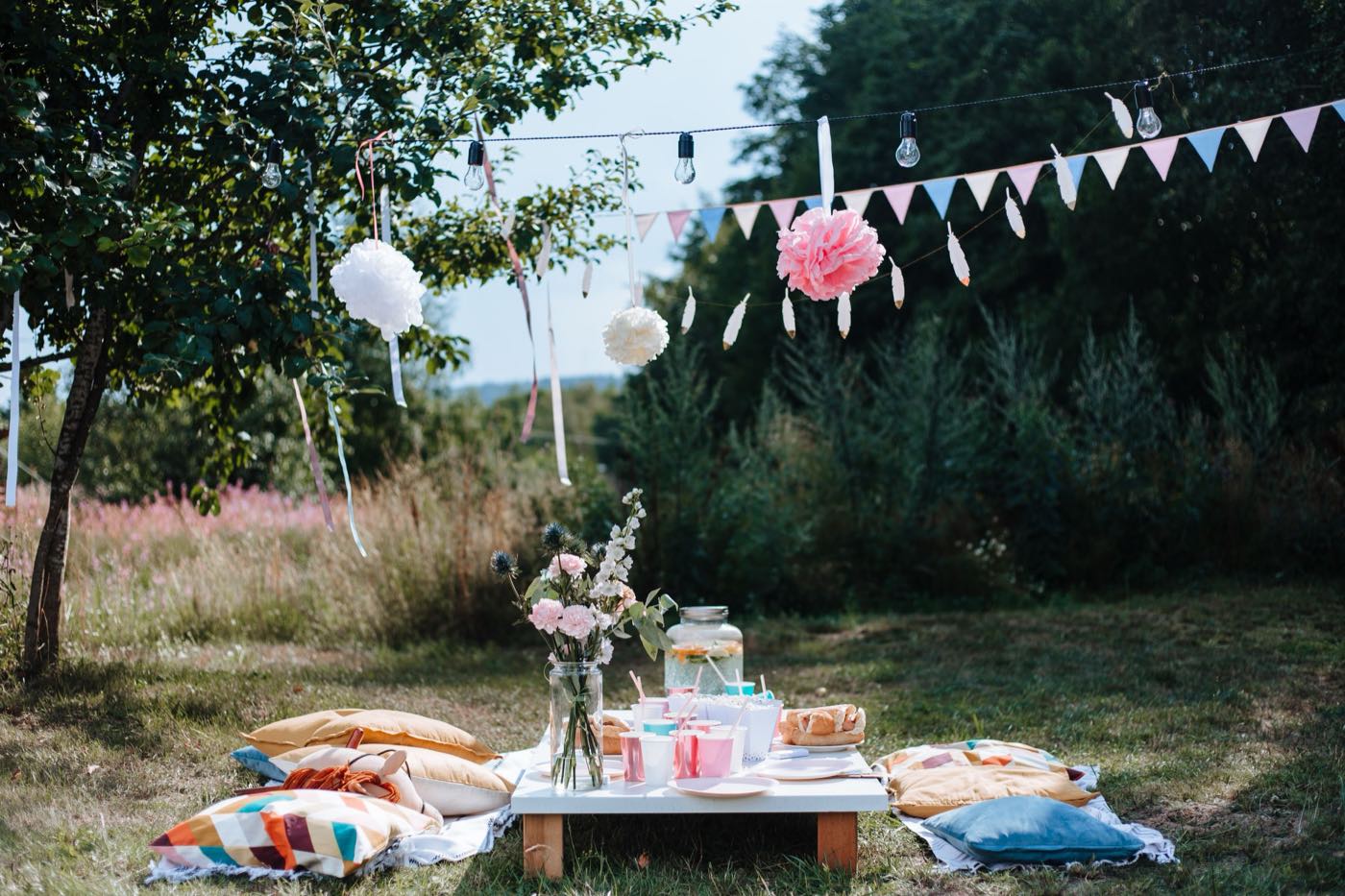 A backyard party on a lawn with a low table of cups and flowers, cushions on the grass, and bunting and paper pom poms overhead