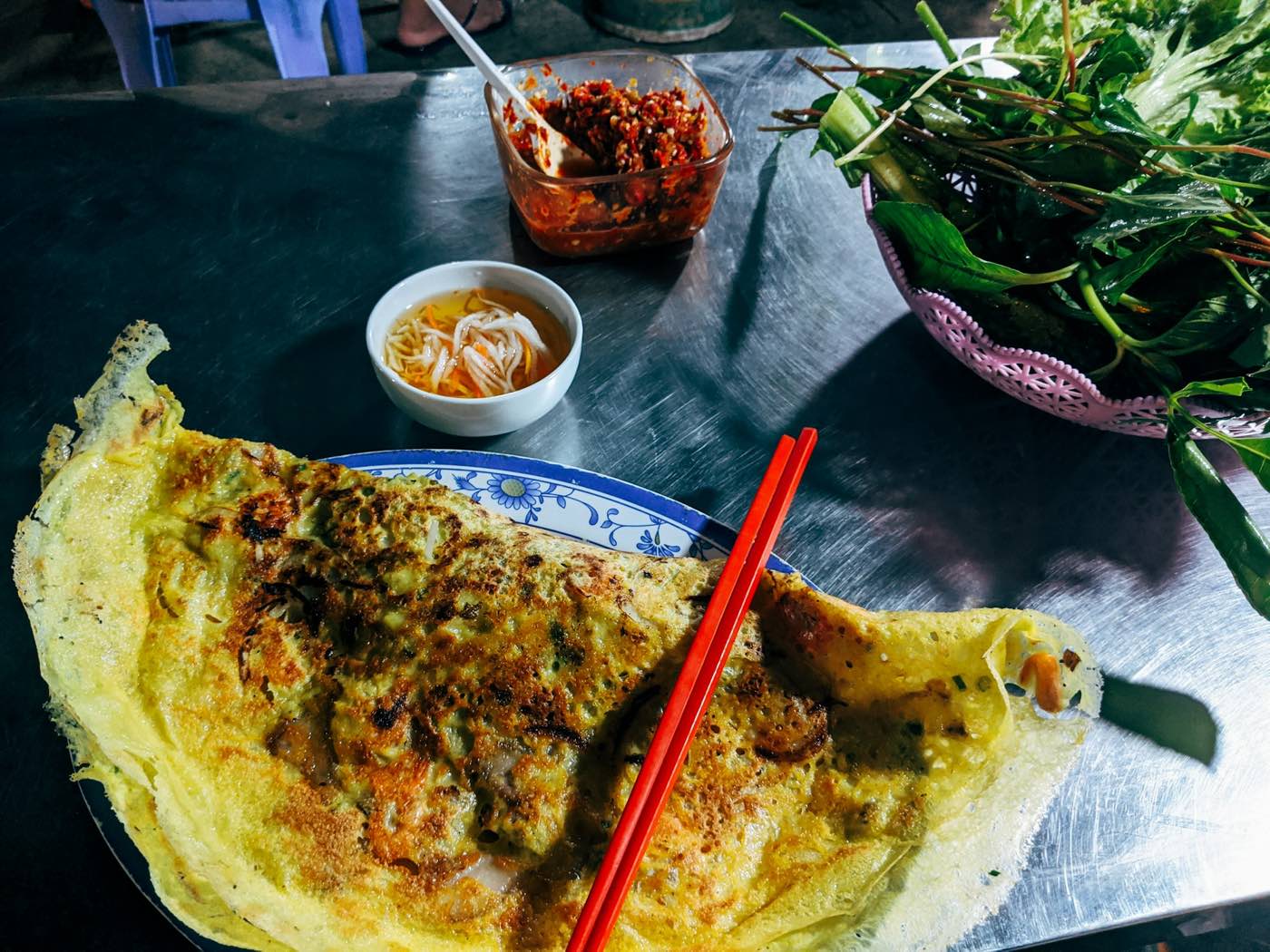A crisp savory crepe on a plate with red chopsticks, dipping sauces and a basket of fresh greens