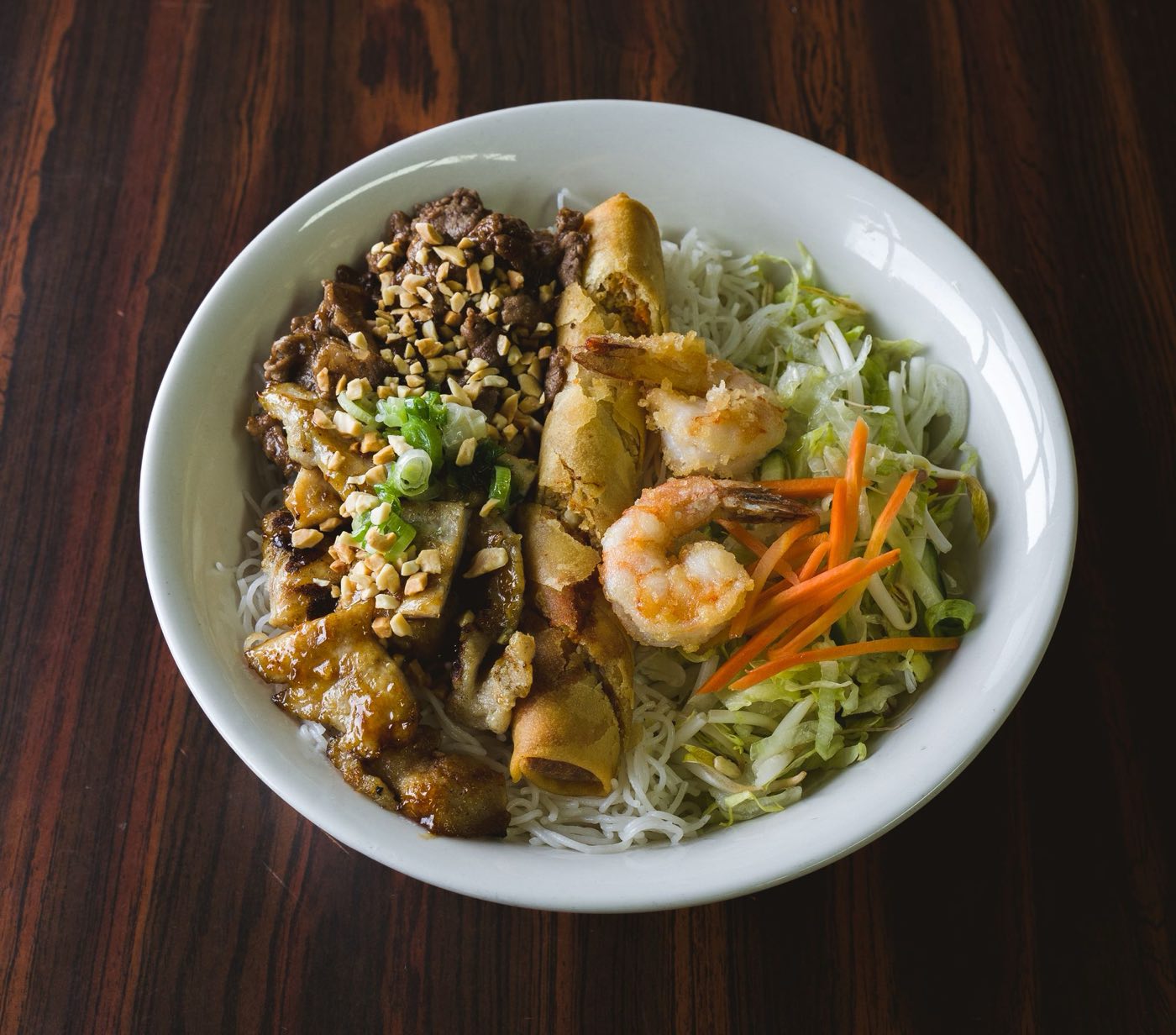 A rice noodle bowl with grilled pork, sliced egg rolls, shrimp, lettuce, carrots and peanuts