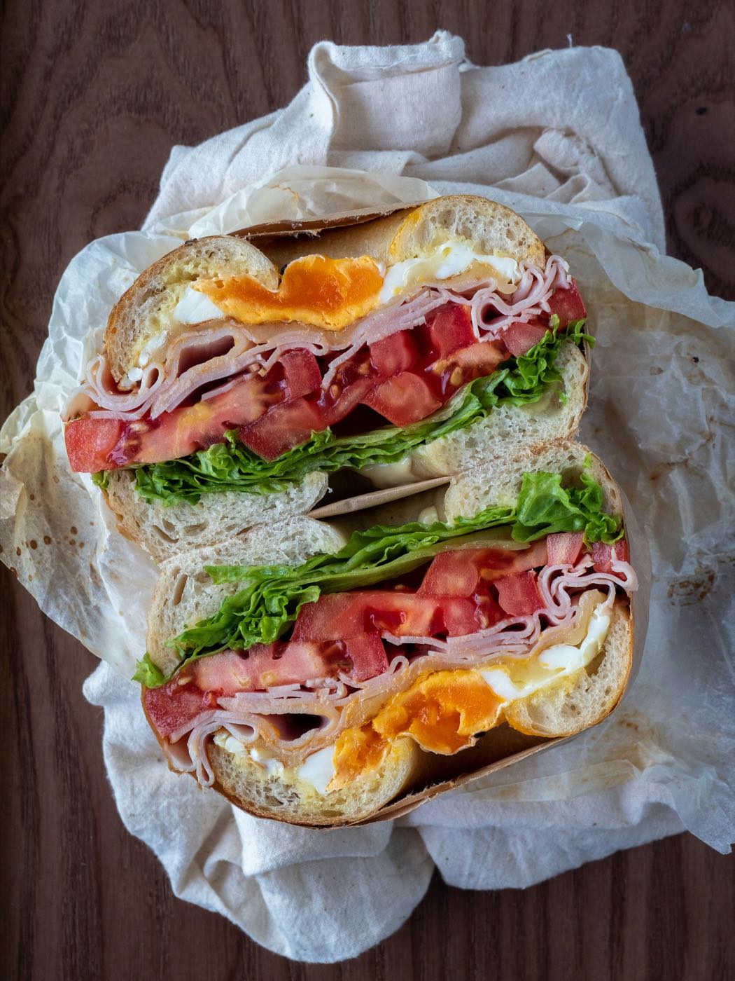 A bagel sandwich with egg, ham, tomato and lettuce, cut in half on white paper