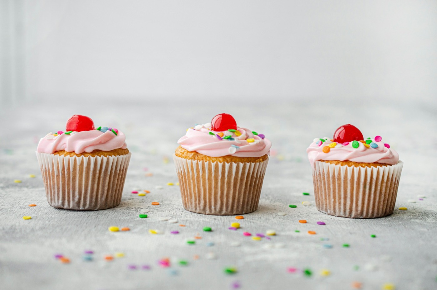 Three cupcakes with pink frosting swirls, red cherries and sprinkles