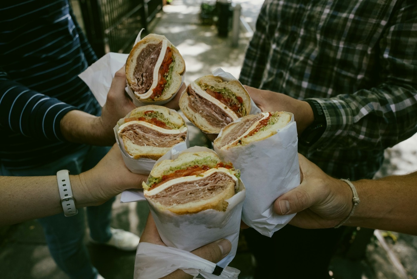 Hands holding halves of sub sandwiches with sliced meats, cheese, tomato and lettuce, wrapped in white deli paper.