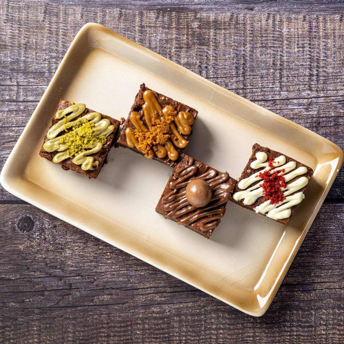 Four brownie squares with drizzled toppings on a cream serving tray.
