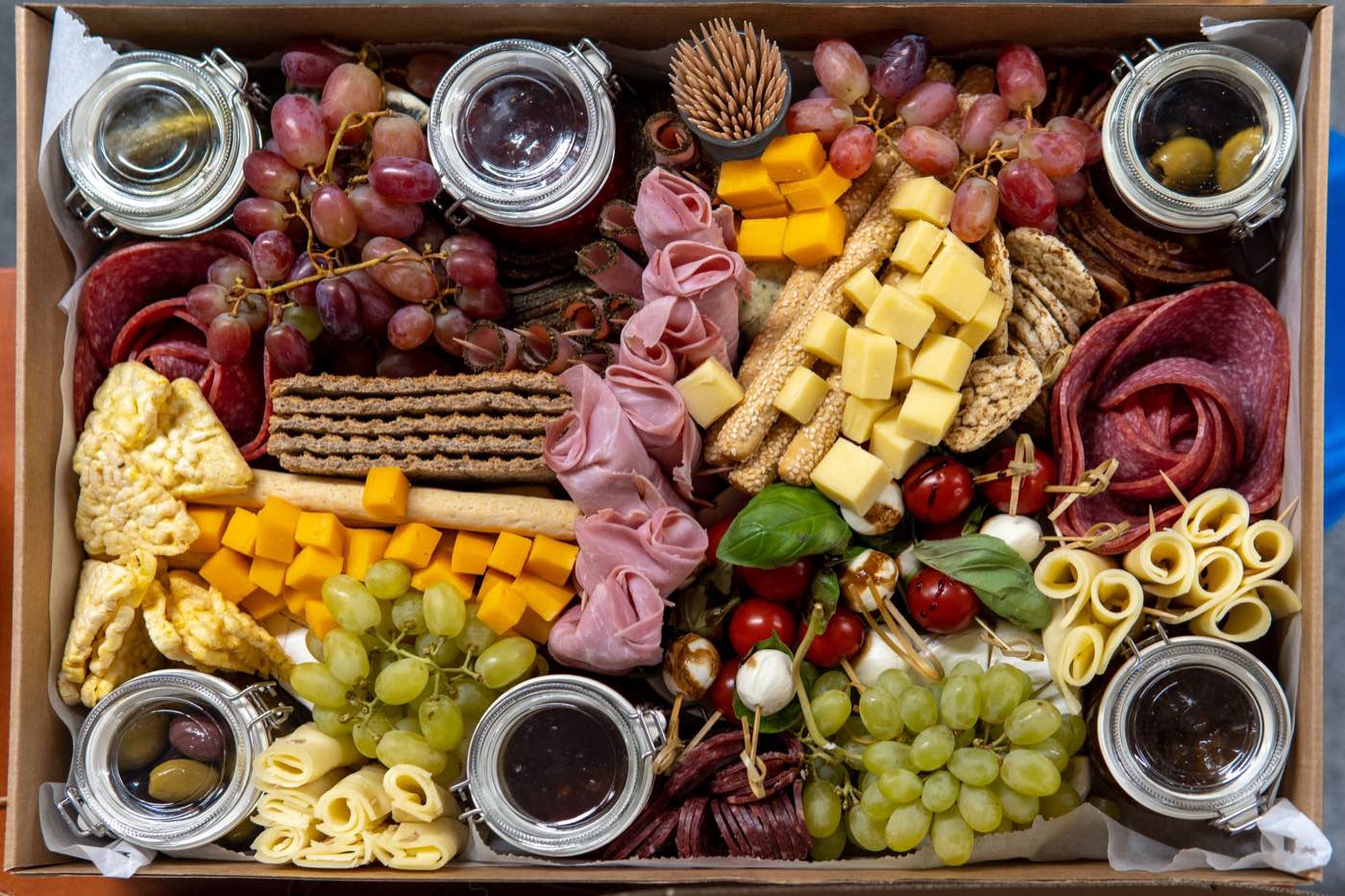 A charcuterie box with salami, rolled ham, cubed cheese, grapes, crackers, tomato and mozzarella skewers, and small jars of olives and jam.