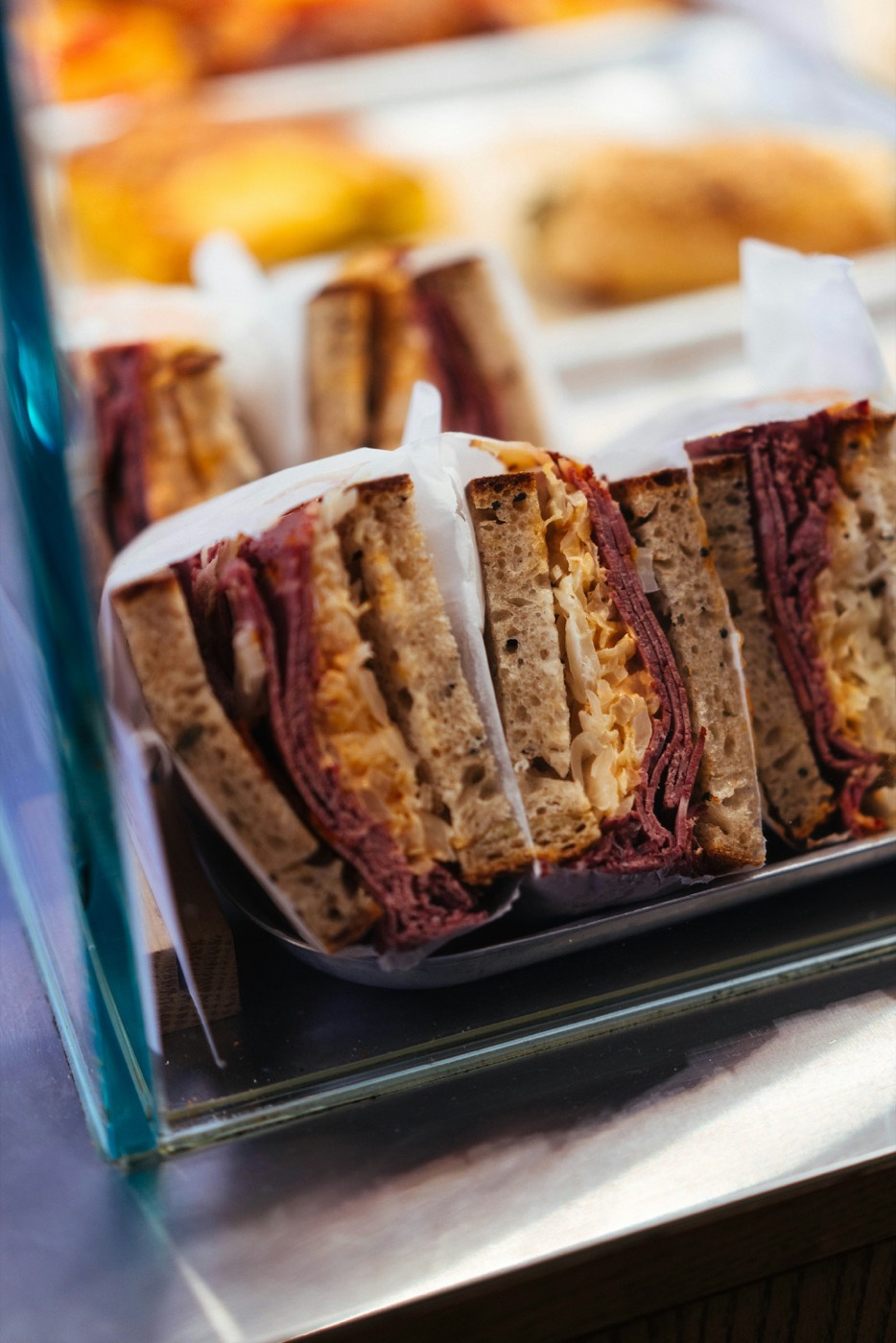 Sandwiches on rye with sliced beef and sauerkraut, cut in half and wrapped in paper, in a glass deli case.