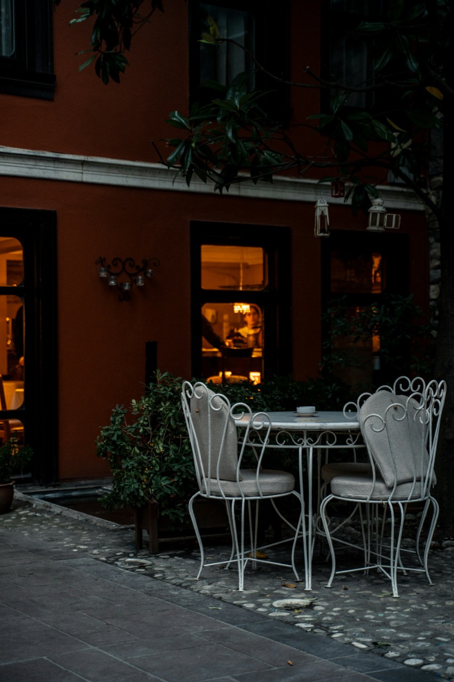 A white iron bistro table and two cushioned chairs on a stone patio outside a lit window at dusk.