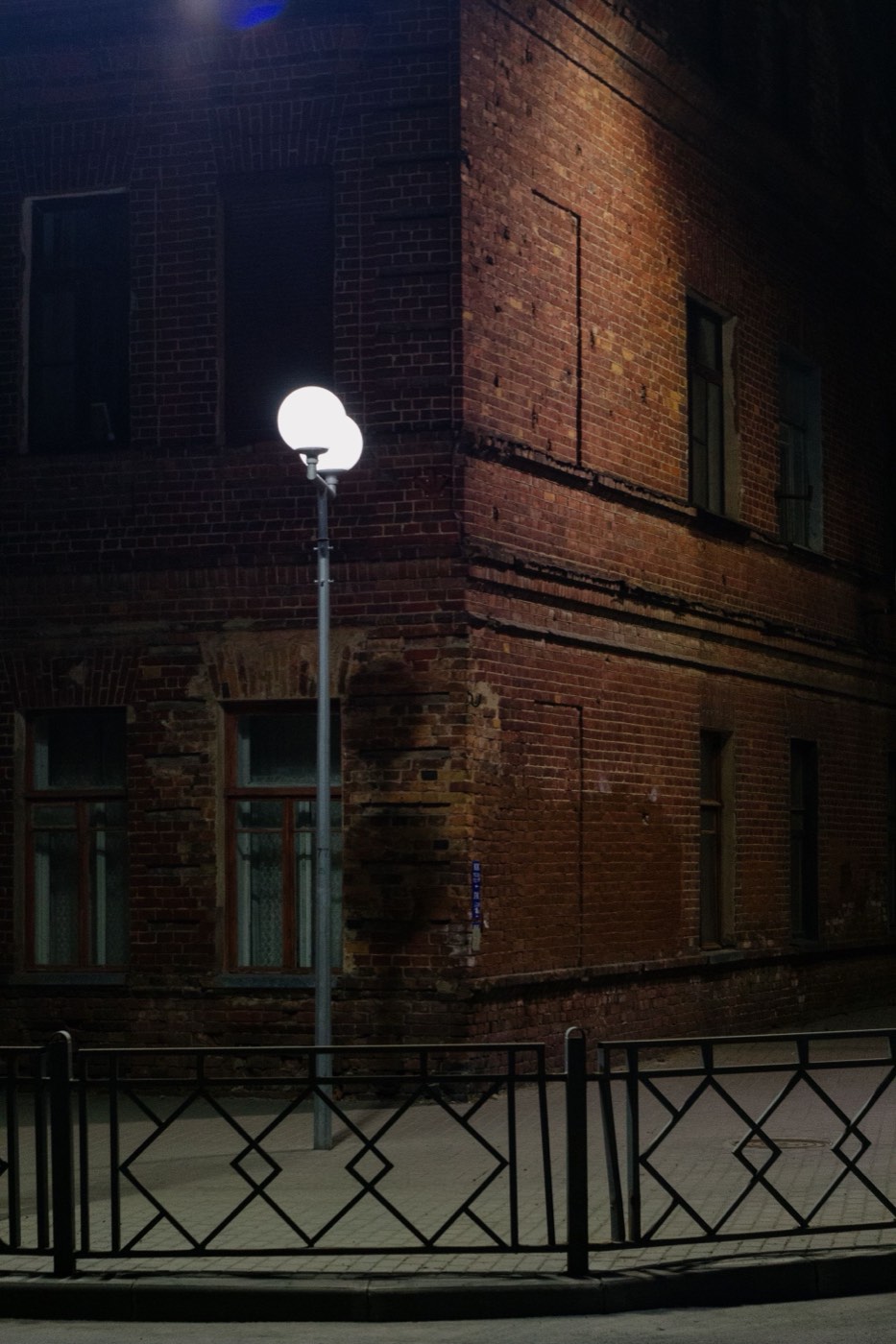 The corner of an old brick building at night, lit by a single street lamp, with an iron fence in front.