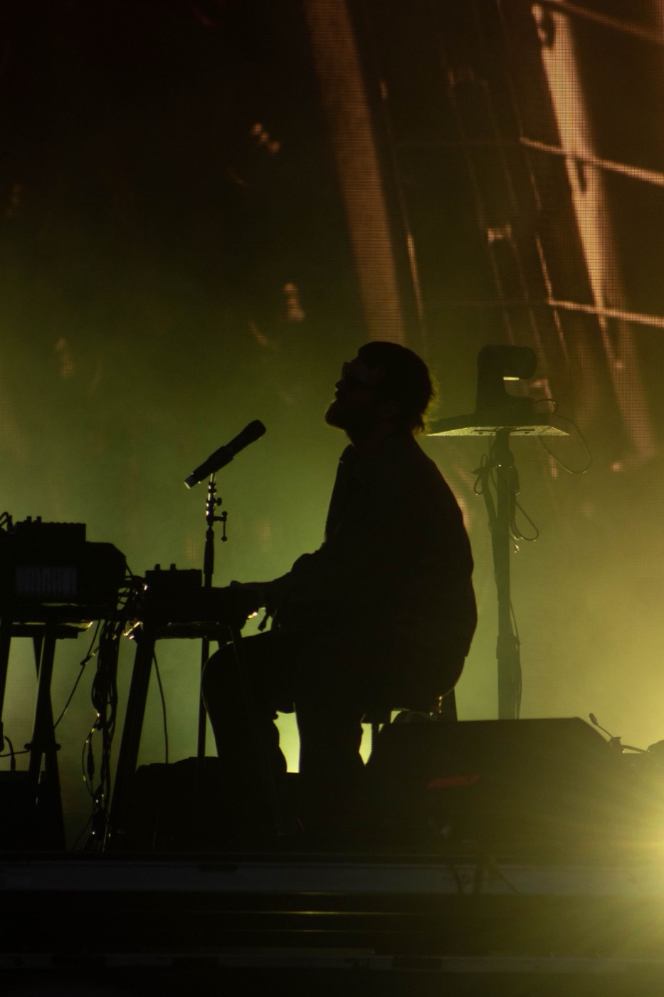 A singer at a keyboard with a microphone, in silhouette on a dim stage.