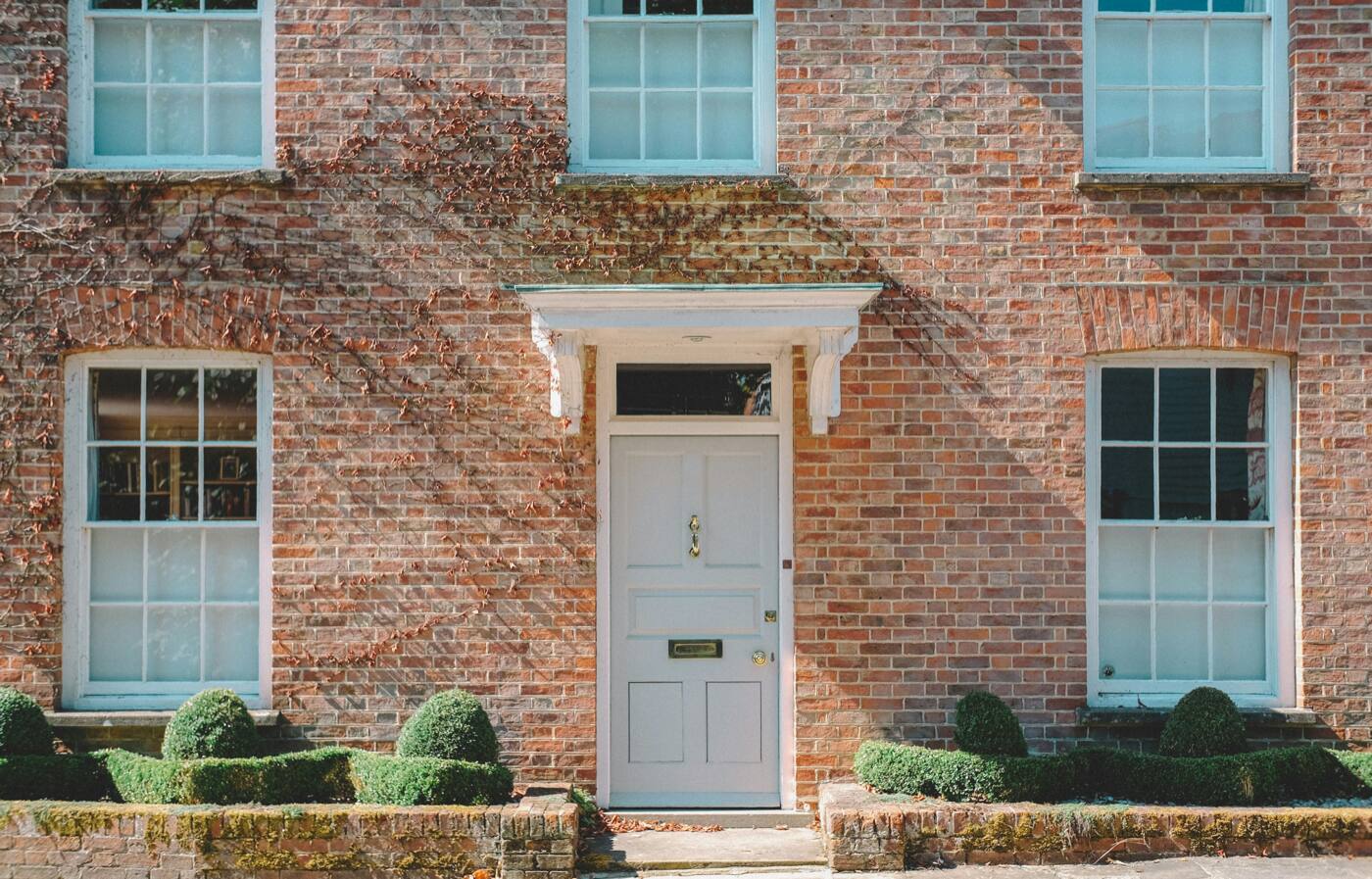 A red brick building with a pale gray front door under a white hood, and trimmed shrubs along the front