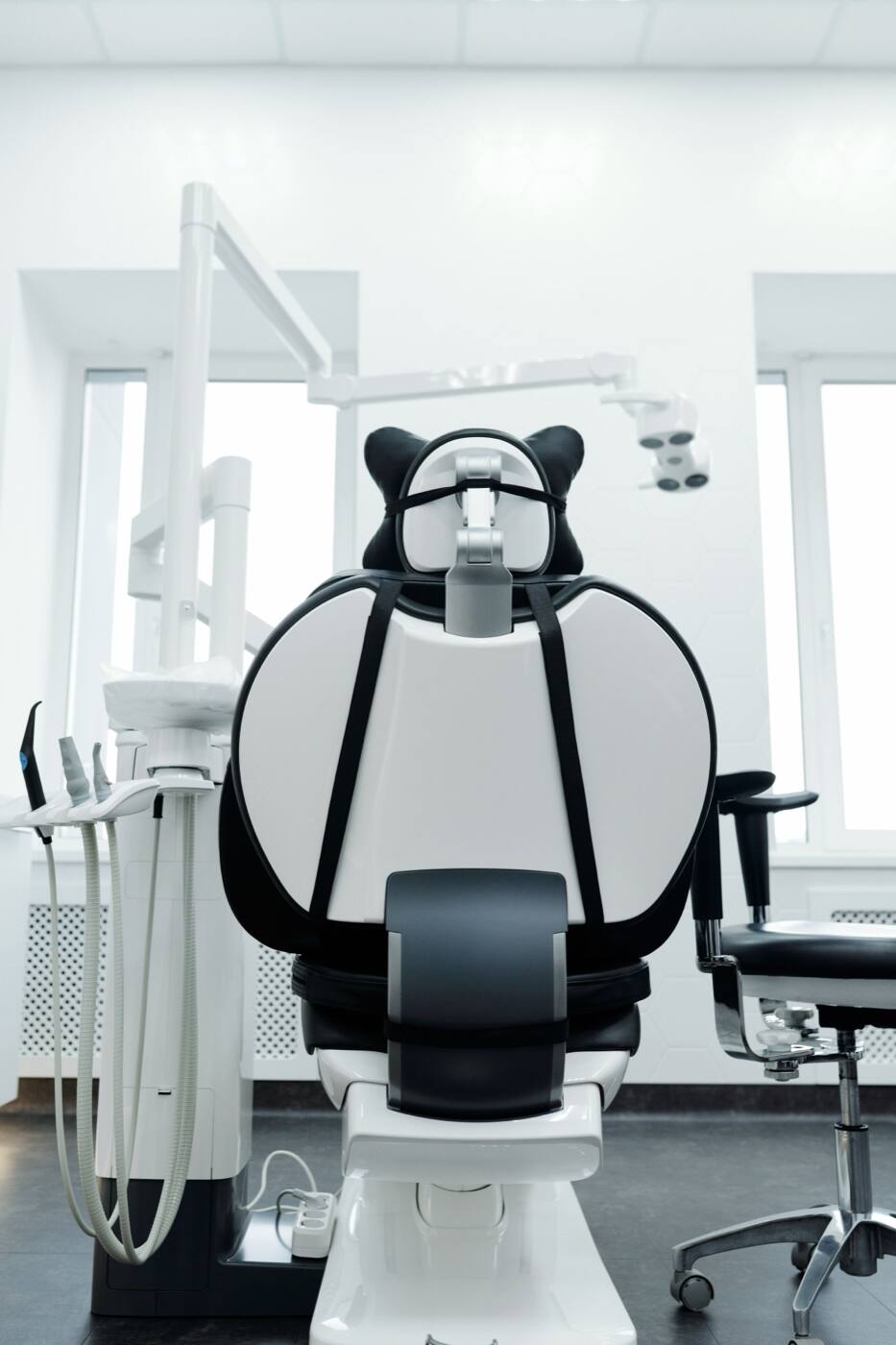 An empty dental chair in a bright white treatment room with two tall windows.