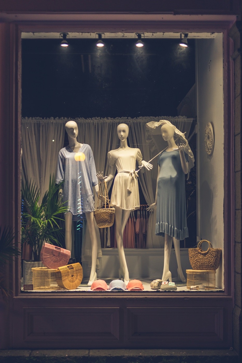 A shop window at dusk with three mannequins in summer dresses, woven bags and caps
