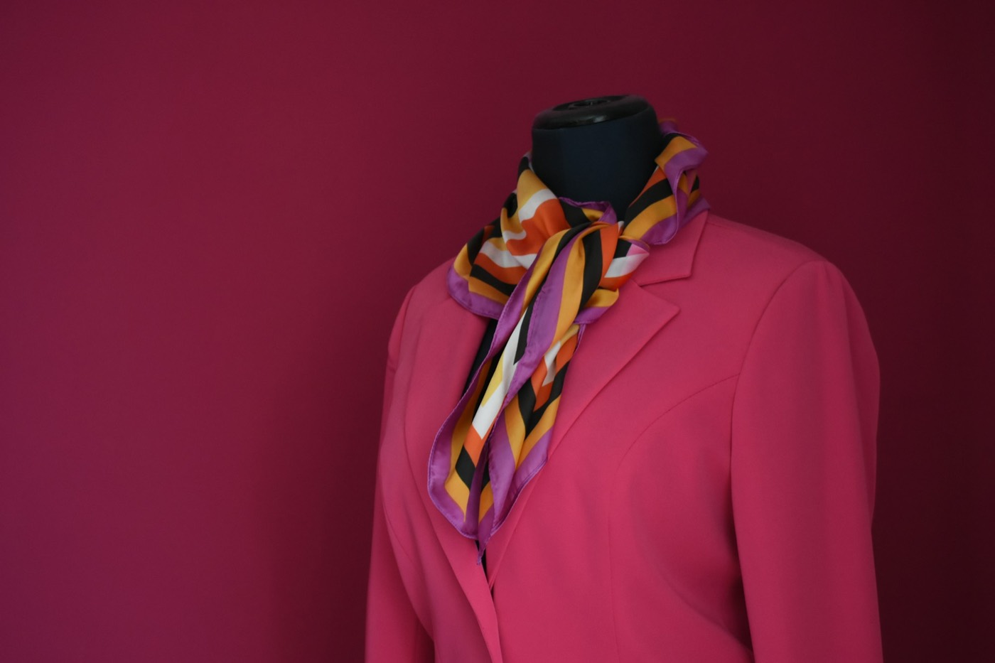 A pink jacket and a striped silk scarf on a dress form against a wine-colored wall