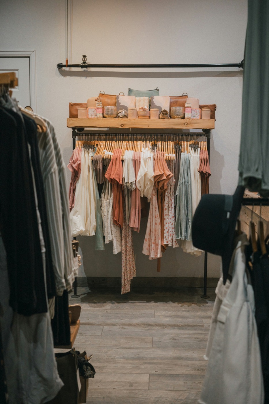 Racks of soft-colored tops and dresses under a wood shelf of handbags and candles