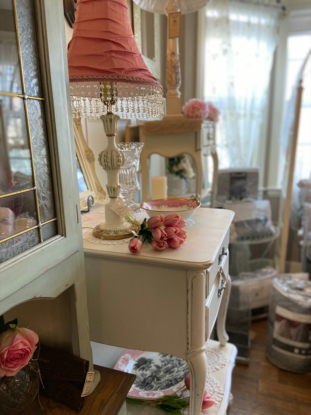 A shop corner with a pale green painted cabinet, pink roses and a fringed pink lamp on a white side table