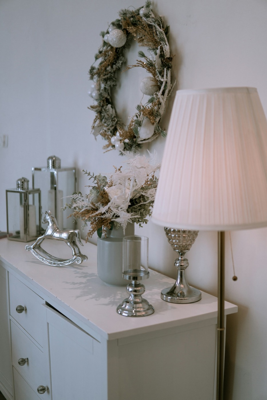 A white dresser styled with a pleated lamp, silver candle holders and a winter wreath