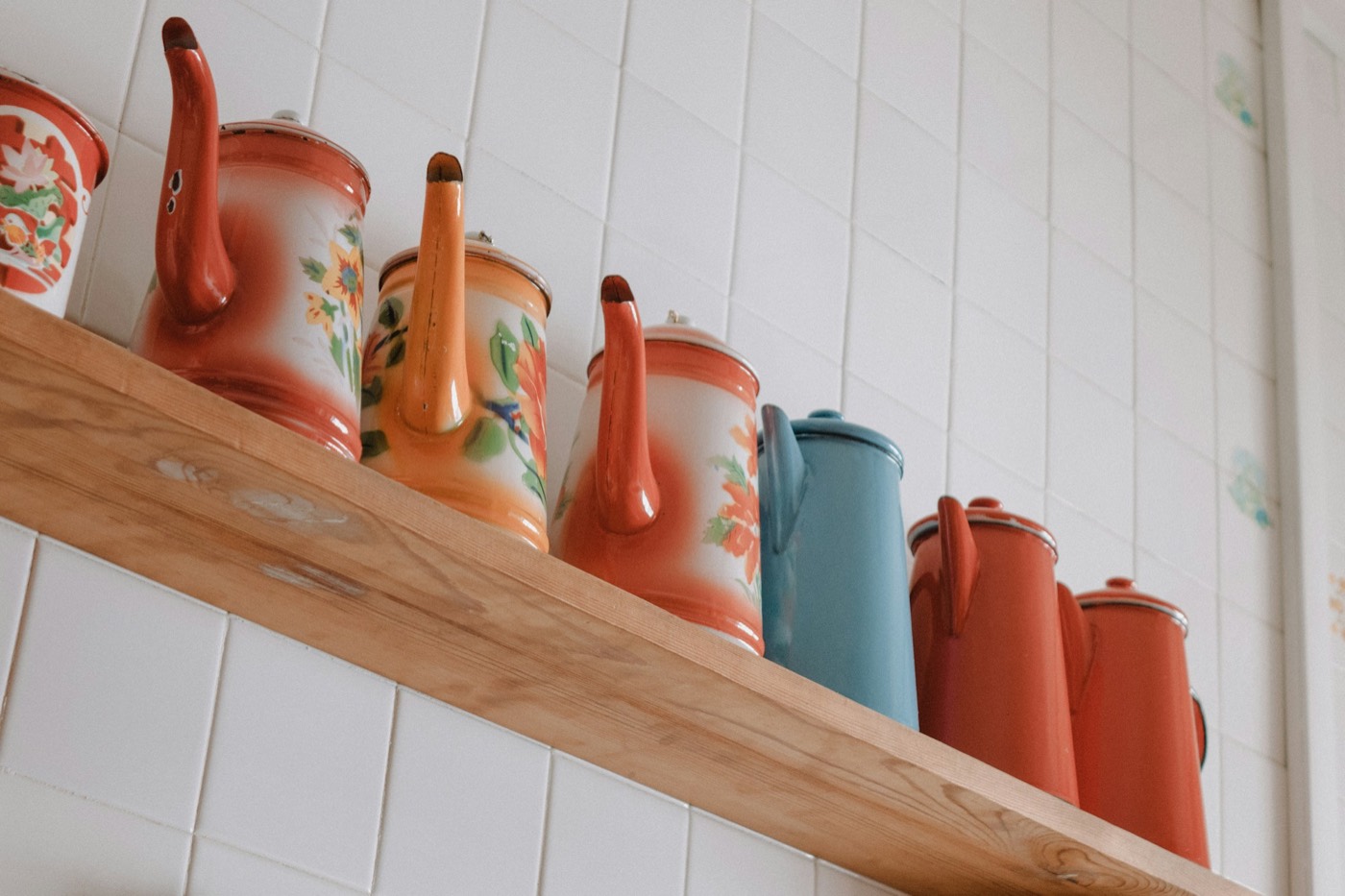Painted enamel coffee pots in red, orange and blue lined up on a wooden shelf