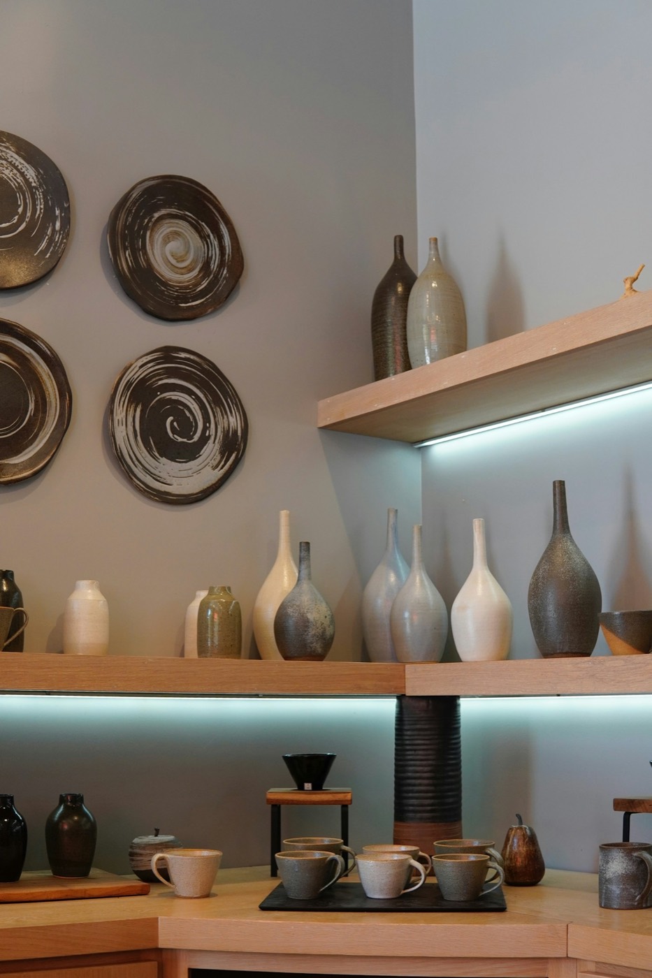 Handmade vases, cups and swirled wall plates on lit wooden shelves