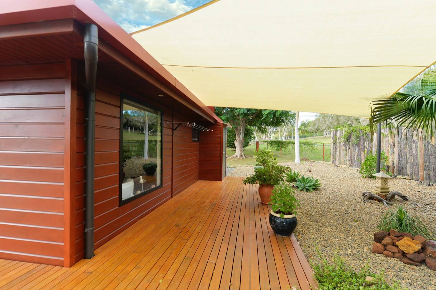 A warm-toned wood deck along a stained wood wall, with a shade sail and a gravel garden.