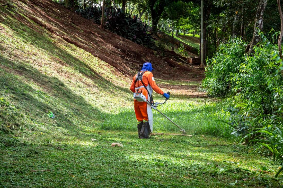 A landscaper in orange hi-vis coveralls trimming grass on a slope, seen from behind.