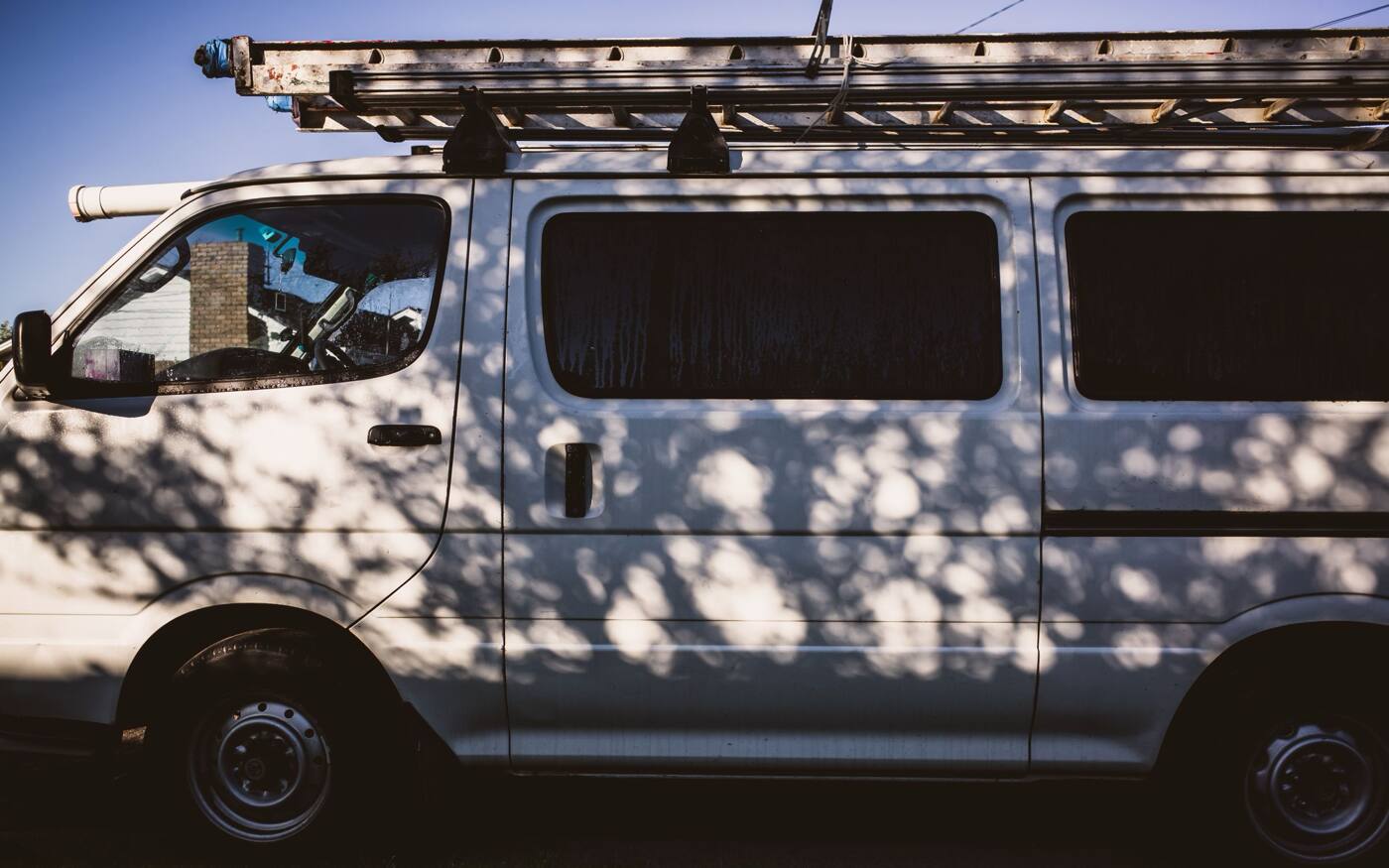 A white work van with a ladder on its roof rack, parked in dappled shade