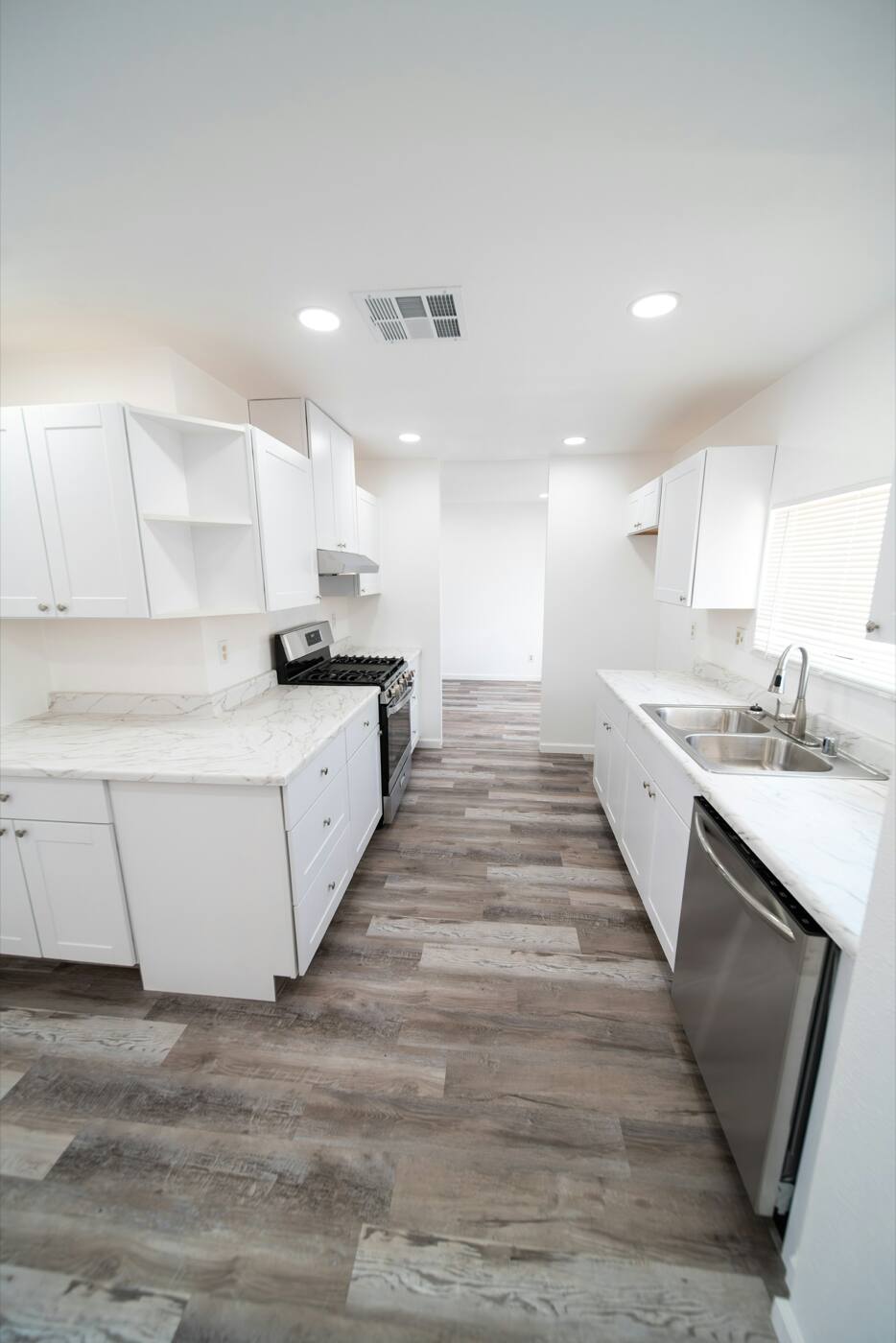 A white kitchen with a ceiling air vent, recessed lights and a gray wood-look plank floor