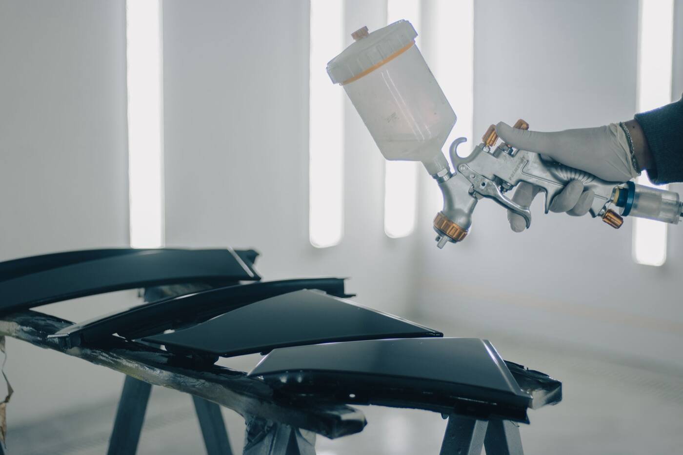 A gloved hand spraying black car panels in a paint booth