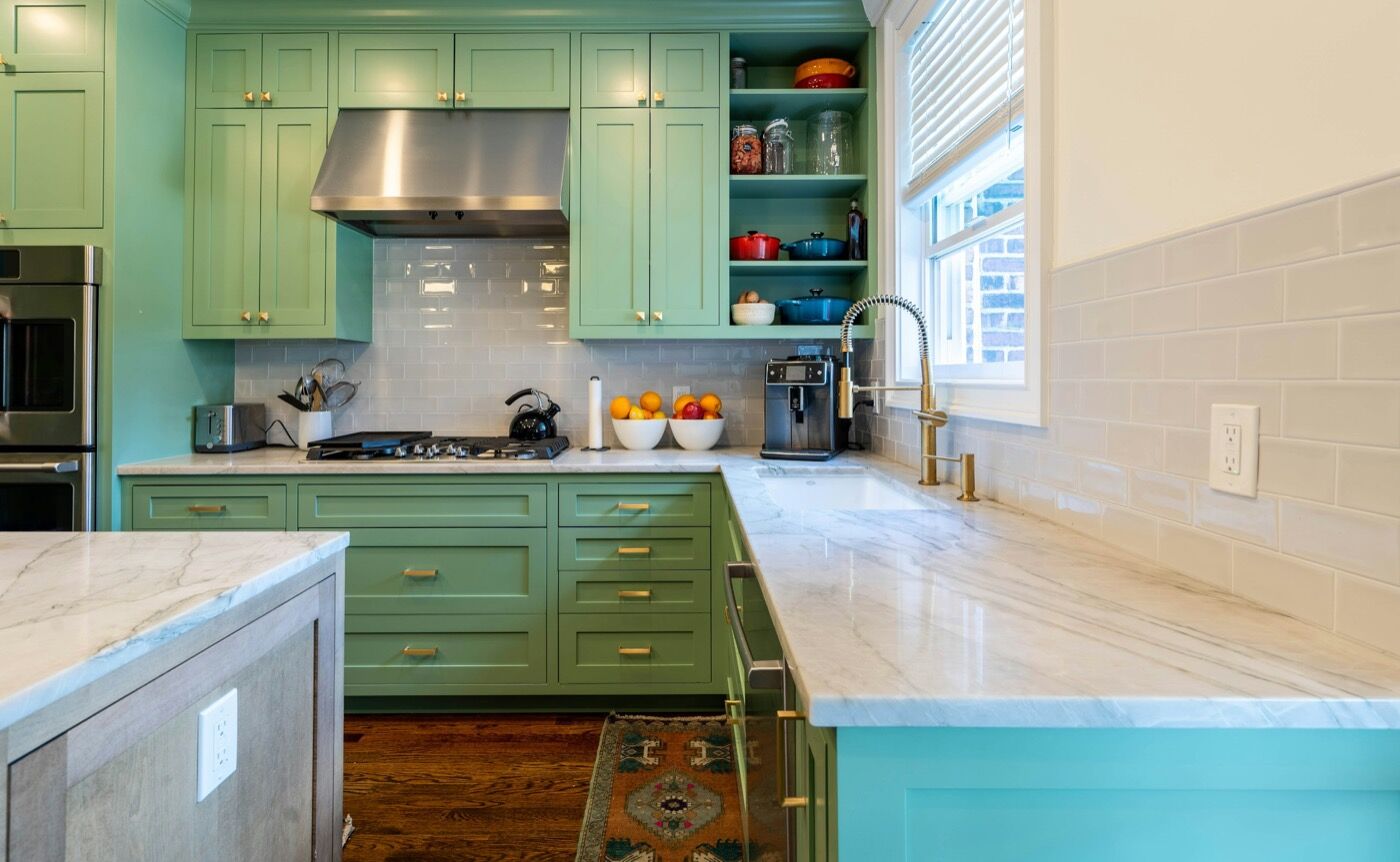 A kitchen with sage green painted cabinets, brass pulls and a white tile backsplash