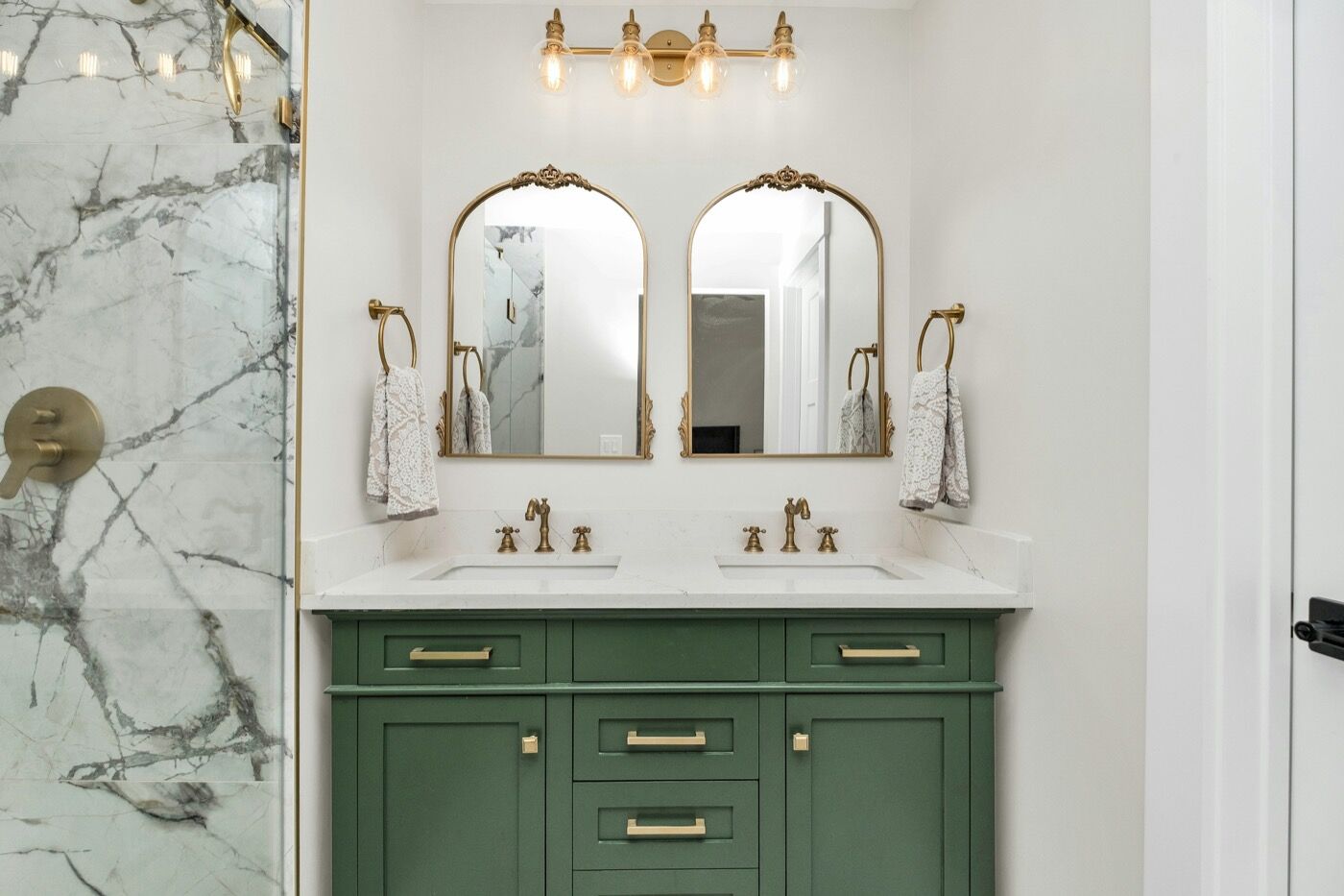 A bathroom with a green painted double vanity and two arched brass mirrors